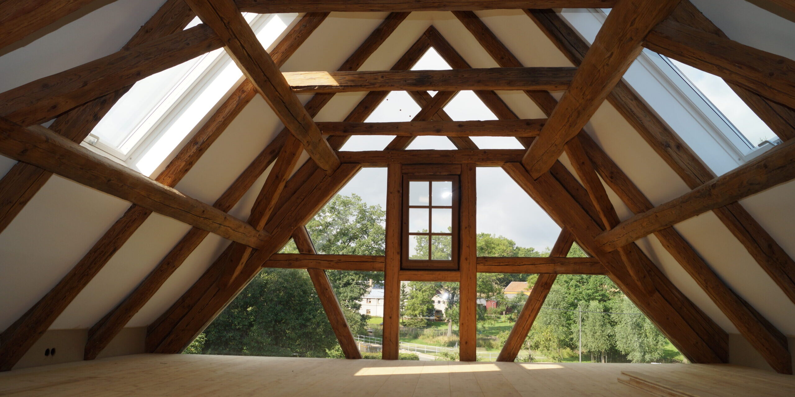 Innenansicht eines hellen Dachgeschosses mit freigelegten, massiven Altholz-Balken einer Fachwerkkonstruktion und einer großzügigen Giebelverglasung.